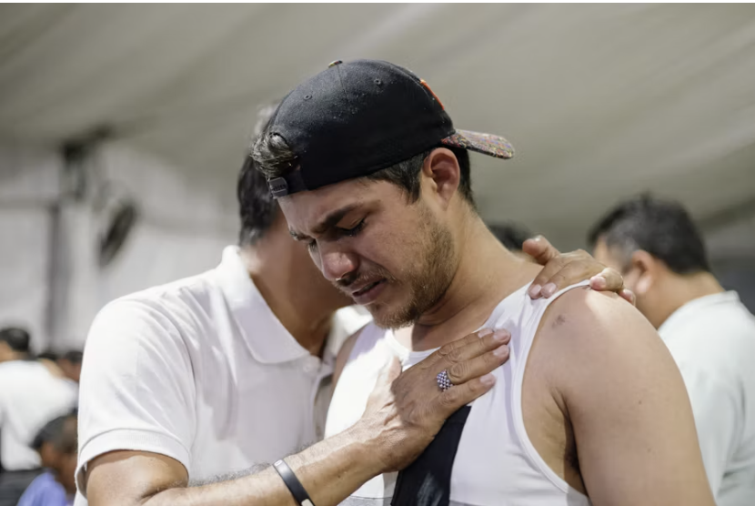 A man rests a hand on his chest while another man supports him with a hand on his shoulder at a community gathering