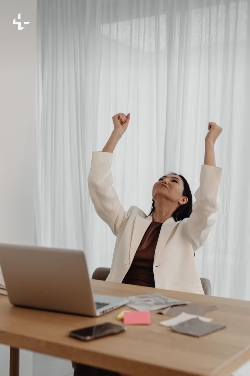 A woman at a bright desk raises both arms in celebration after finishing a piece of work