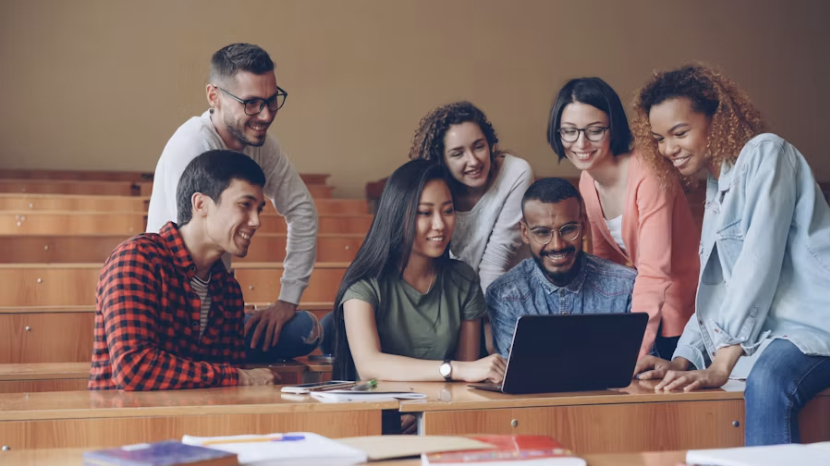 A group of students and colleagues gather around a laptop in a lecture hall, smiling as they review training material together