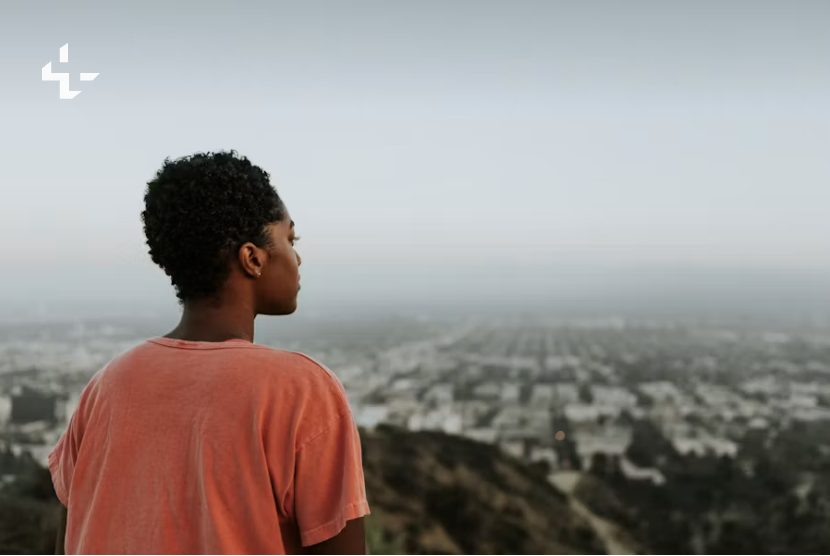 A young person seen from behind looks out over a hazy city skyline from a hilltop