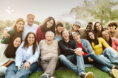 A large multigenerational group sits together on the grass in a park, smiling at the camera