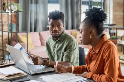 A mentor and a colleague review notes together at a laptop in a warm shared workspace