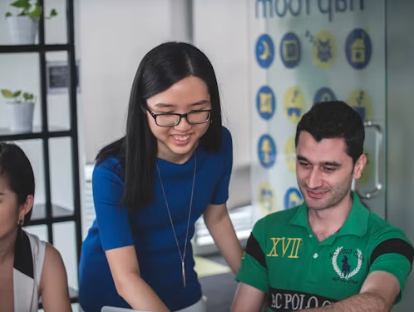 A team member leans over a desk to review work on a laptop with two smiling colleagues