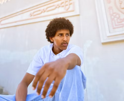 A young man in a white shirt sits outdoors against a pale wall, reaching one hand forward