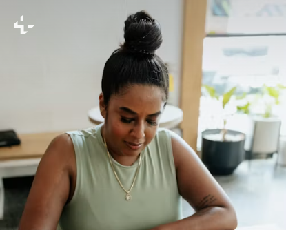 A woman in a sage green top works with focus at a bright table in a sunlit office